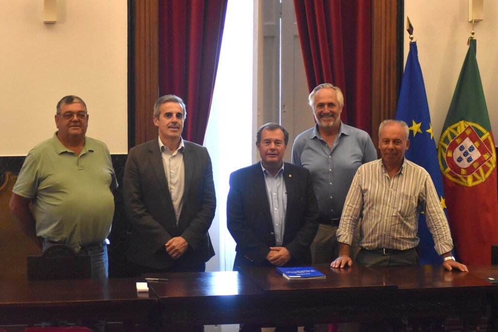 Cinco homens em pé junto a uma mesa durante a assinatura de um protocolo, com as bandeiras de Portugal e da União Europeia ao fundo.
