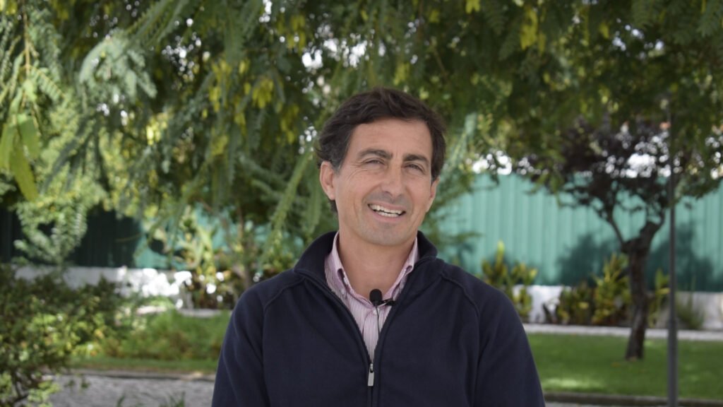 Homem de meia-idade sentado ao ar livre, sorridente, com camisa clara e casaco azul escuro, em frente a árvores e vegetação num espaço ajardinado.