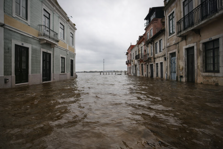 Rua da zona ribeirinha inundada devido à subida do caudal do rio Tejo, com água a atingir a entrada dos edifícios.
