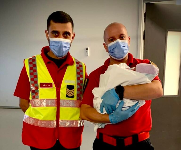 Dois bombeiros fardados seguram um recém-nascido ao colo no interior de uma unidade de saúde.