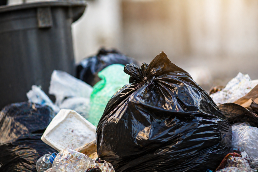 Sacos de lixo e resíduos acumulados junto a um contentor, ilustrando problemas de deposição e limpeza urbana