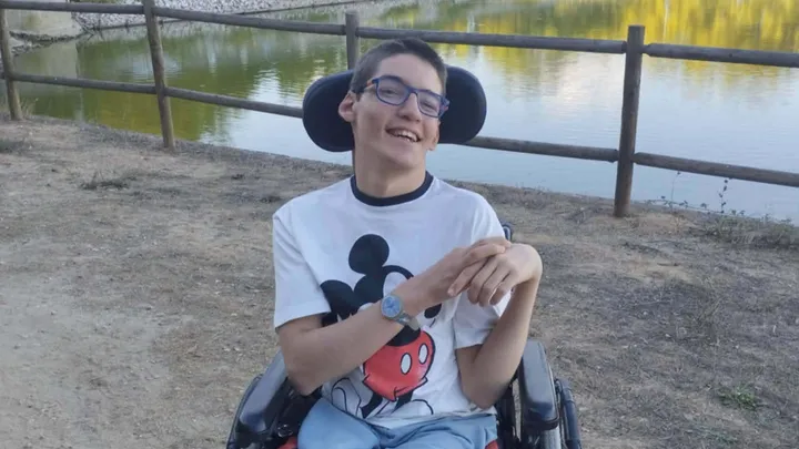 Jovem de 19 anos em cadeira de rodas, sorridente, ao ar livre junto a um lago