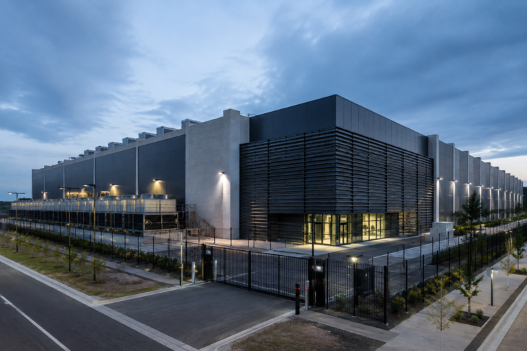 Vista exterior de um data center moderno, com edifício industrial de grande dimensão, vedação de segurança e equipamentos técnicos de refrigeração. Imagem ilustrativa.