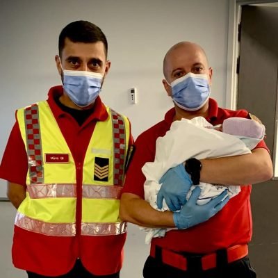 Dois bombeiros fardados seguram um recém-nascido ao colo no interior de uma unidade de saúde.