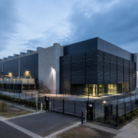 Vista exterior de um data center moderno, com edifício industrial de grande dimensão, vedação de segurança e equipamentos técnicos de refrigeração. Imagem ilustrativa.