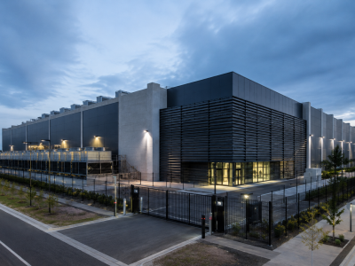 Vista exterior de um data center moderno, com edifício industrial de grande dimensão, vedação de segurança e equipamentos técnicos de refrigeração. Imagem ilustrativa.