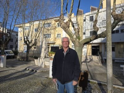 Homem de pé numa praça na Castanheira do Ribatejo, em frente a um edifício institucional, com árvores sem folhas ao redor, num dia de céu limpo.