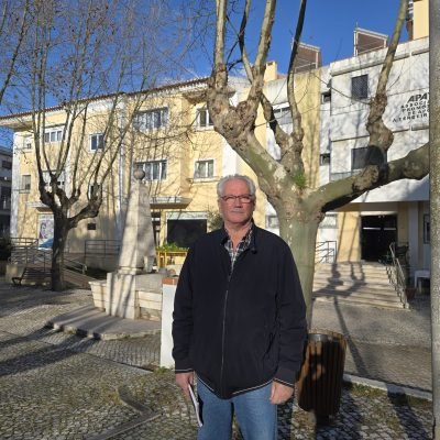 Homem de pé numa praça na Castanheira do Ribatejo, em frente a um edifício institucional, com árvores sem folhas ao redor, num dia de céu limpo.